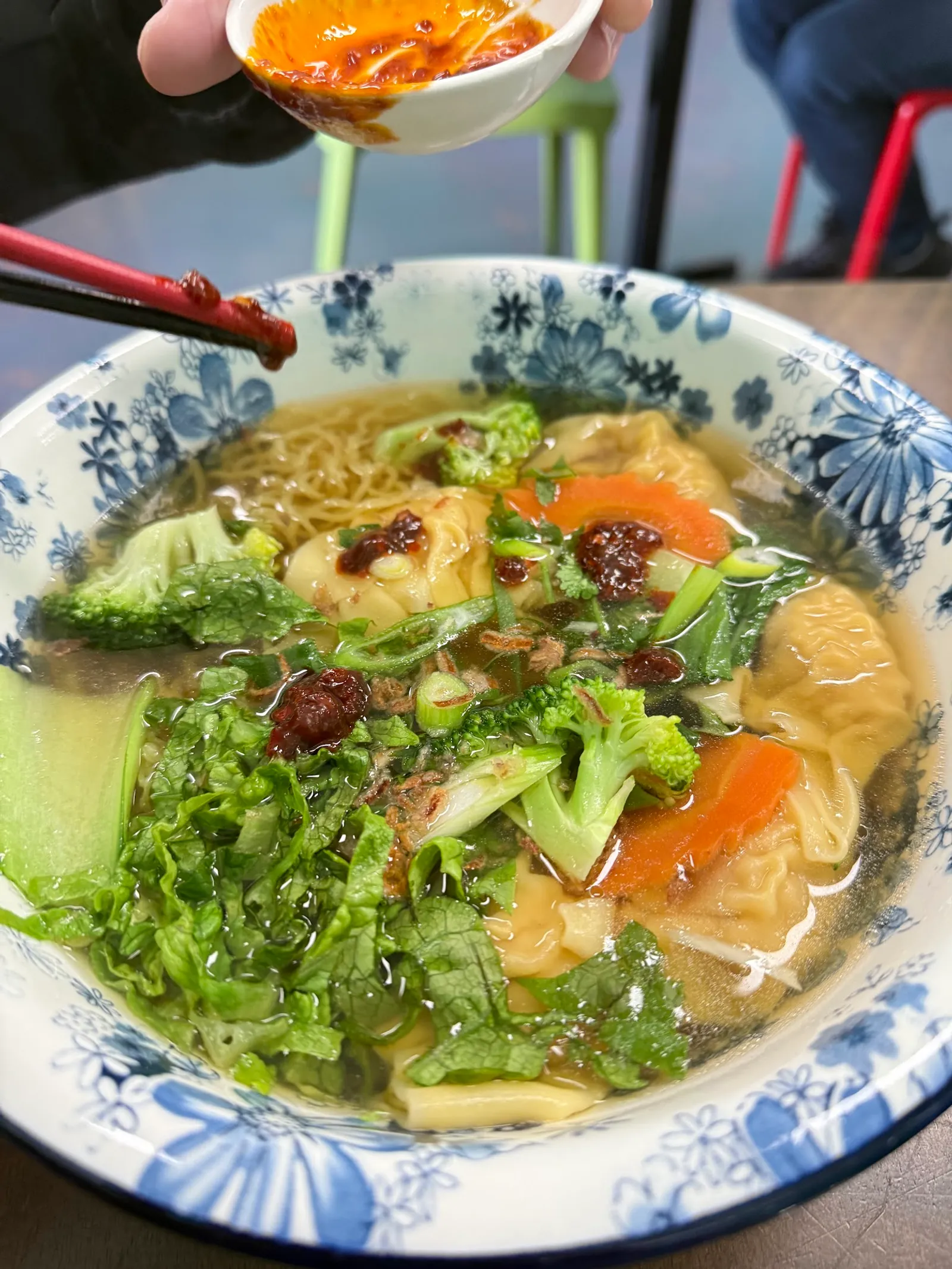 Wonton noodle soup in a blue-and-white bowl with a hand adding chili sauce
