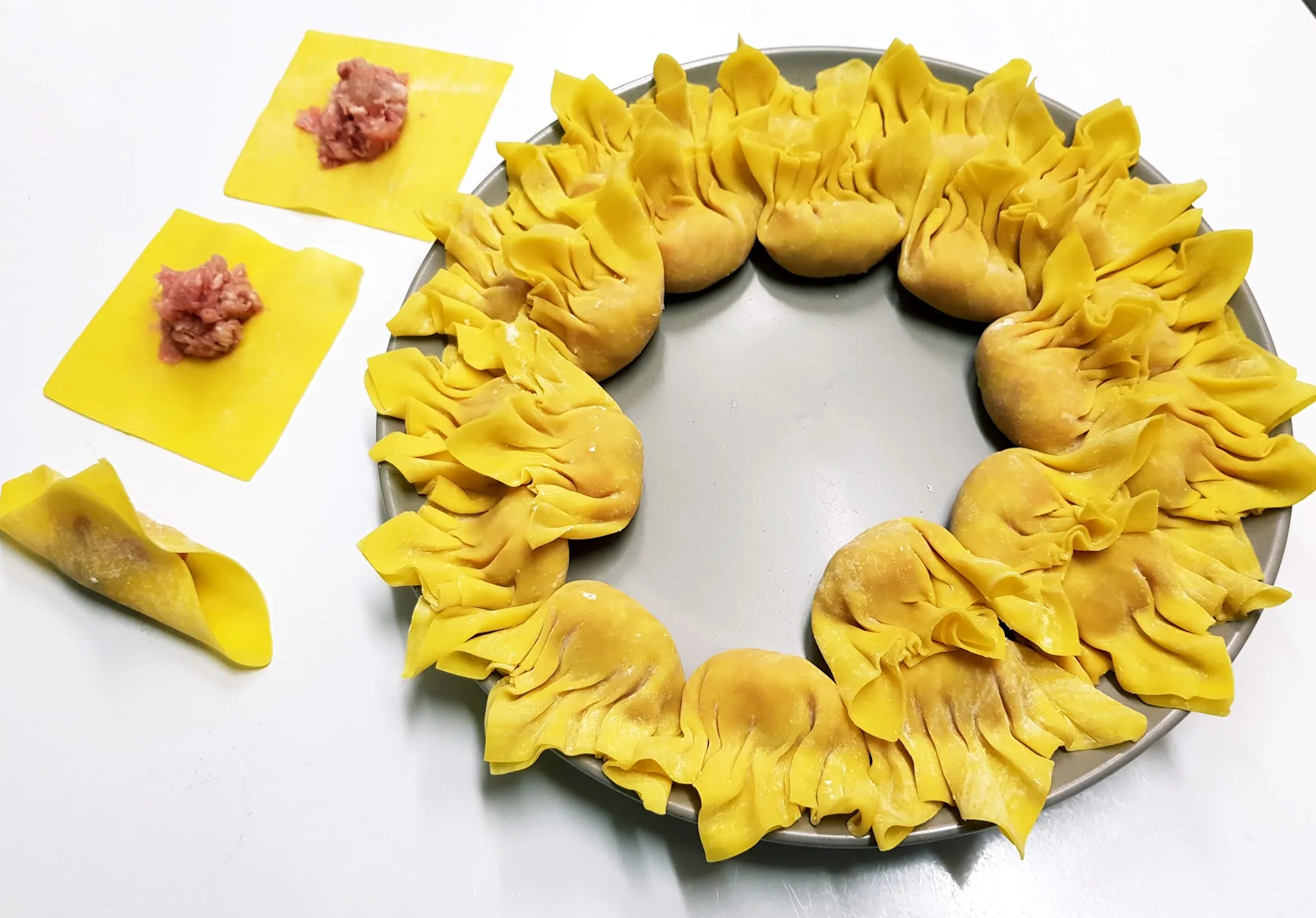Hand-made wontons arranged in a circle on a tray with loose wrappers and filling beside them